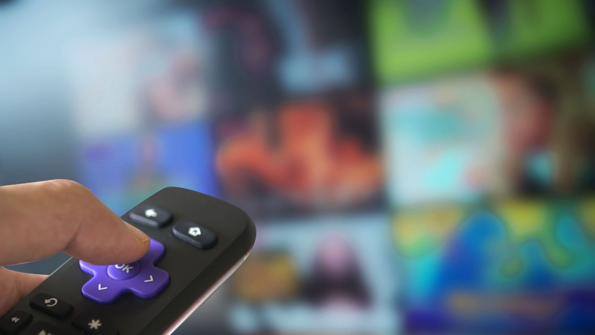 Hand holding TV remote with blurry television screen in background