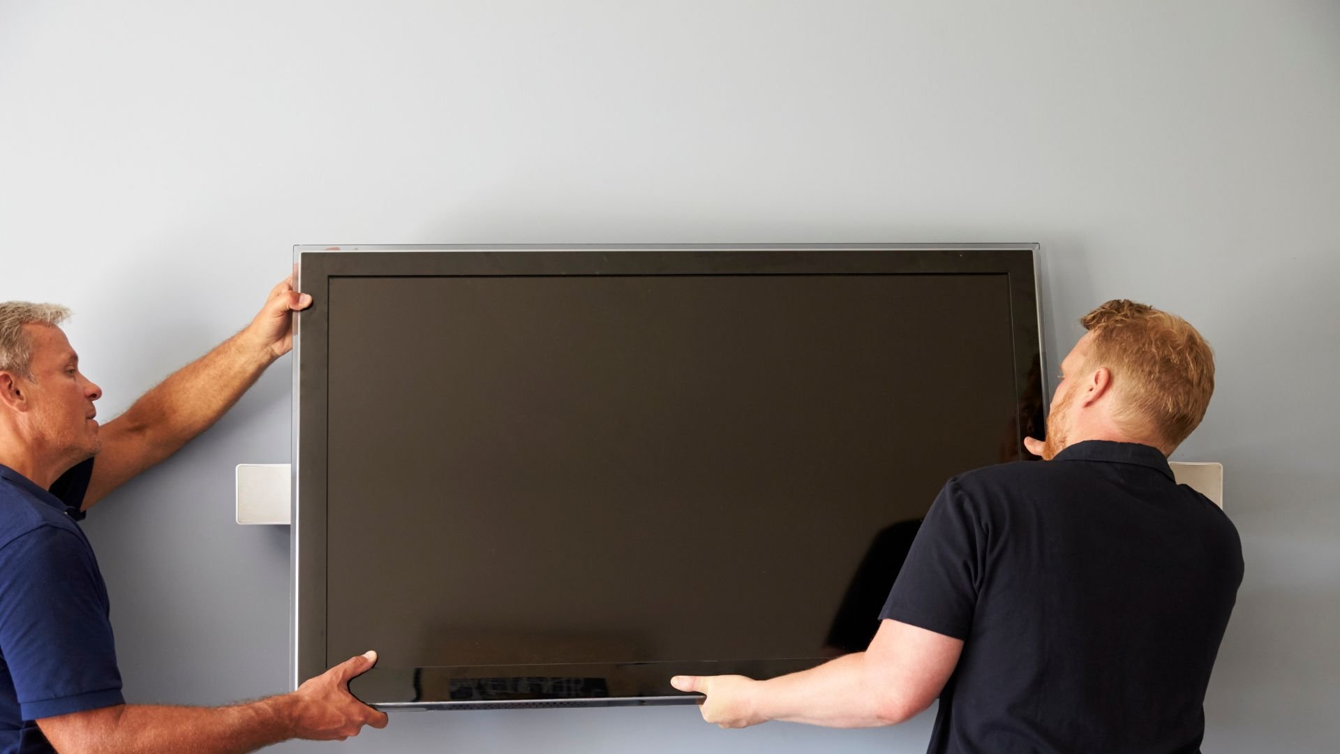 Two men carefully mounting a large black flat-screen television on wall